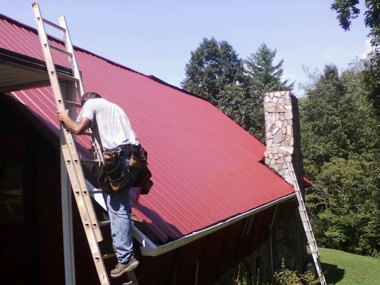 Roofer on a ladder beside a red metal roof and stone chimney — Soffit & Fascia Repair by Roofing Brothers in Plumerville